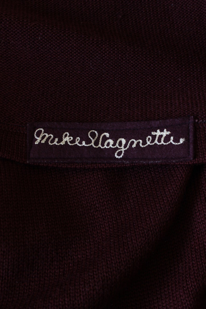 May include: Close-up of a dark burgundy knit fabric with a sewn-on label. The label is a darker shade of burgundy and features the embroidered text "Mike Magnetti" in white thread. The fabric appears to be a sweater or similar garment.