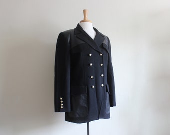 May include: A black double-breasted blazer with leather accents on the shoulders and pockets. The blazer features gold-colored buttons and is displayed on a mannequin. A classic and stylish outerwear garment.