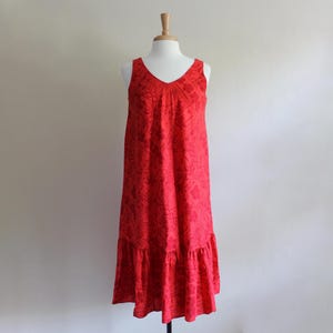 May include: A sleeveless midi dress in a vibrant red hue, featuring a V-neckline and a subtle, tonal floral pattern throughout the fabric. The garment has a relaxed, A-line silhouette that finishes with a gathered ruffle hem at the bottom.