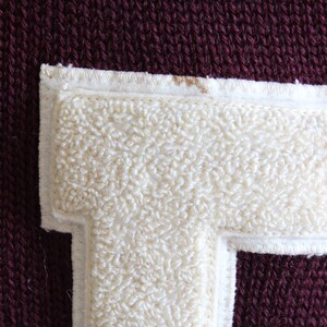 May include: Close-up of a maroon knit fabric with a cream-colored, fuzzy letter "T" patch. The patch is outlined with a white border and stitched with white thread. The knit fabric has a textured appearance.