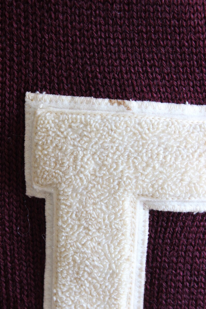 May include: Close-up of a maroon knit fabric with a cream-colored, fuzzy letter "T" patch. The patch is outlined with a white border and stitched with white thread. The knit fabric has a textured appearance.