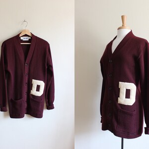 May include: Maroon cardigan sweater with a large cream-colored "D" patch on the front. The sweater has a V-neck, button closure, and two front pockets. The sleeves and bottom hem have ribbed cuffs.