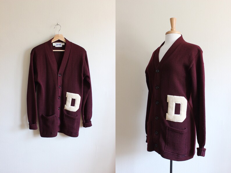 May include: Maroon cardigan sweater with a large cream-colored "D" patch on the front. The sweater has a V-neck, button closure, and two front pockets. The sleeves and bottom hem have ribbed cuffs.