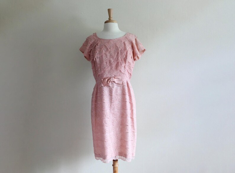 May include: A pale pink knee-length dress featuring an all-over floral lace overlay, short ruffled sleeves, a round neckline, and a matching satin bow detail centered at the waistline. The garment is displayed on a mannequin against a plain wall.