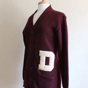 May include: A maroon cardigan sweater with a cream-colored letter "D" patch. The sweater has a V-neck, button closure, and two front pockets. The cuffs and hem are ribbed.