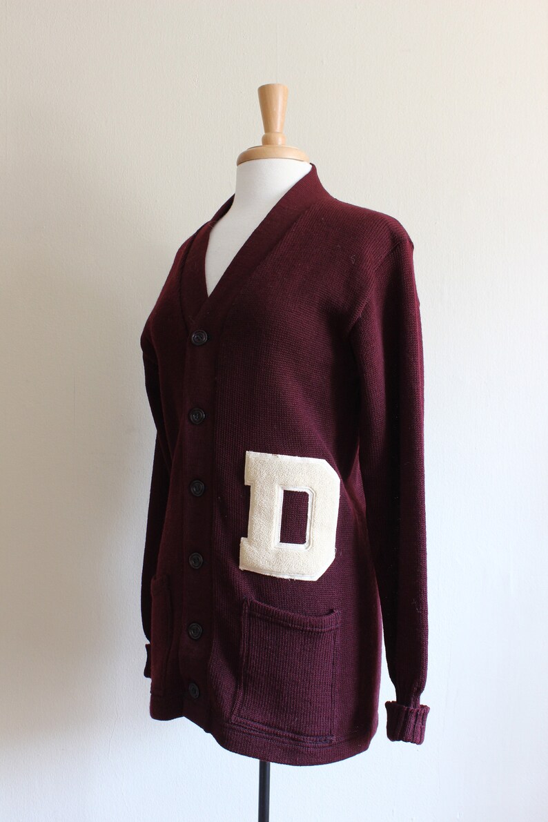 May include: A maroon cardigan sweater with a cream-colored letter "D" patch. The sweater has a V-neck, button closure, and two front pockets. The cuffs and hem are ribbed.