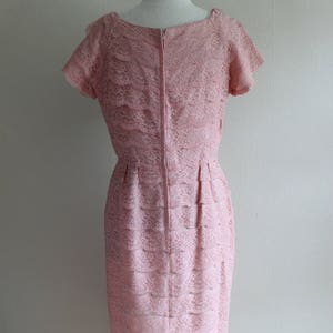 May include: A light pink dress featuring a layered lace overlay with a floral pattern. The garment has short sleeves, a round neckline, and a vertical center back zipper closure. The skirt portion displays horizontal tiers of lace fabric throughout.
