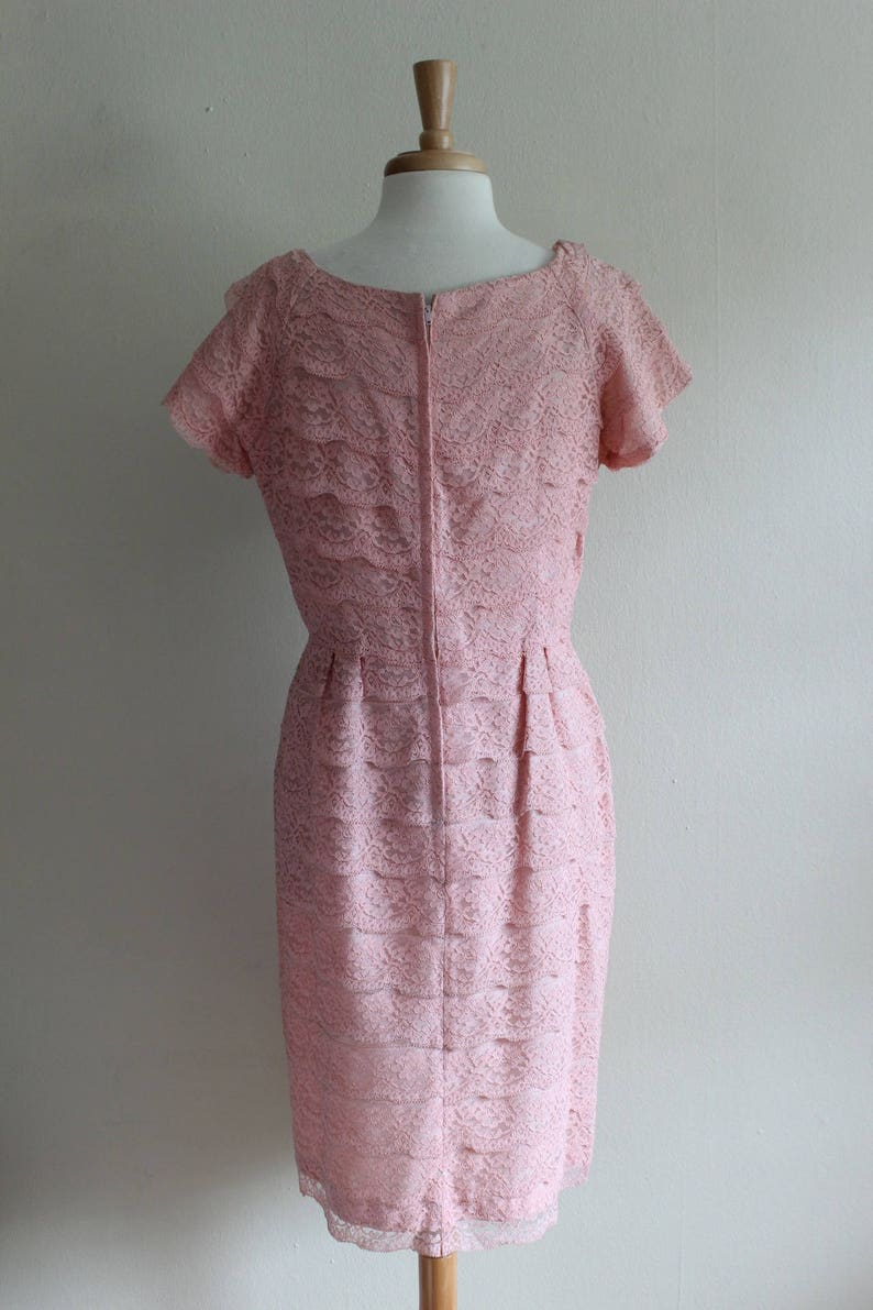 May include: A light pink dress featuring a layered lace overlay with a floral pattern. The garment has short sleeves, a round neckline, and a vertical center back zipper closure. The skirt portion displays horizontal tiers of lace fabric throughout.