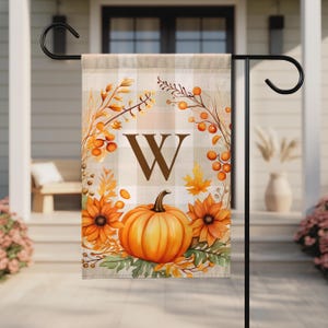 May include: A rectangular garden flag featuring a beige and white buffalo plaid background. The center displays a large brown letter W. Below the letter is a bright orange pumpkin surrounded by orange sunflowers, green leaves, and berry sprigs.