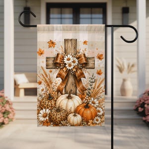 May include: A rectangular garden flag featuring a wooden cross adorned with a large brown ribbon and white daisies. Below the cross are several pumpkins, including two with leopard print patterns, surrounded by wheat stalks and autumn leaves.