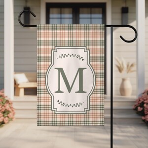 May include: A rectangular garden flag featuring a plaid pattern in muted shades of sage green and soft peach. Centered is a white rectangular frame with decorative corners, containing a large, dark green letter M and small leaf sprig accents.