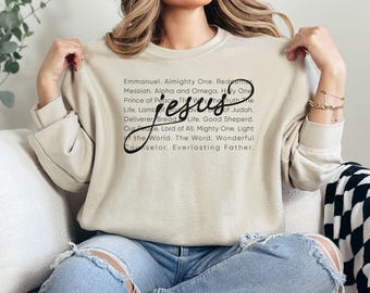 May include: A beige sweatshirt with the word "Jesus" in a cursive font. The sweatshirt has a list of religious terms printed in black. The person is wearing blue jeans.