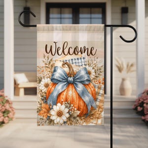 May include: A rectangular garden flag featuring a beige and white plaid background with the word Welcome in dark brown script. An orange pumpkin sits in the center, adorned with a large blue bow, surrounded by white daisies and autumn foliage.