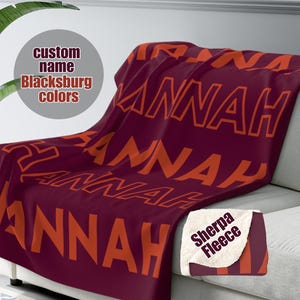 May include: A maroon and orange sherpa fleece blanket with the name "HANNAH" repeated in a bold orange font. The blanket is folded and draped over a couch.