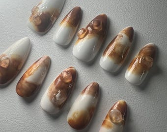 May include: A set of ten almond-shaped false nails. Each nail features a white base with a gradient of brown and amber tones. Some nails have a small, clear, gem-like embellishment near the cuticle.