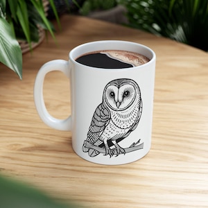 May include: White ceramic coffee mug featuring a black and white illustration of an owl perched on a branch. The mug is filled with dark coffee. The mug has a curved handle and is sitting on a wooden surface.