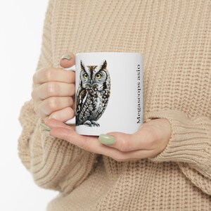 May include: White ceramic mug featuring a detailed illustration of an owl with brown, gray, and white feathers and yellow eyes. The text "Megascops asio" is printed vertically on the side of the mug. The mug is held by a person.