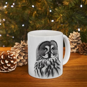 May include: White ceramic mug featuring a detailed black and white illustration of a Great Grey Owl. The owl's face and chest are prominently displayed. The mug has a curved handle and is set against a background of pine cones and greenery.