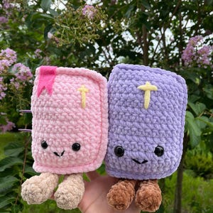 May include: Two plush book-shaped toys made of soft yarn. One is light pink with a pink bookmark and beige legs, the other is light purple with brown legs. Both feature black safety eyes, a stitched smile, and a small yellow cross on the cover.