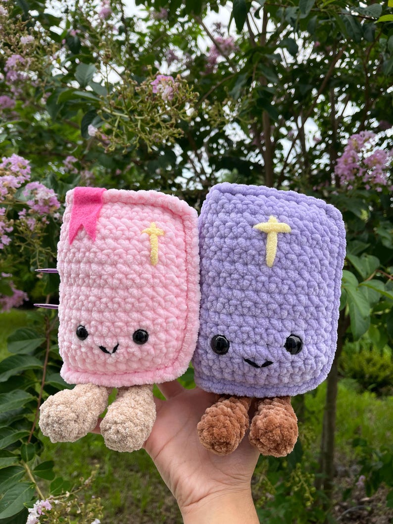 May include: Two plush book-shaped toys made of soft yarn. One is light pink with a pink bookmark and beige legs, the other is light purple with brown legs. Both feature black safety eyes, a stitched smile, and a small yellow cross on the cover.