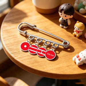 May include: A large silver safety pin with four red circular charms. Each charm has a white design, with the last charm reading "JUNG KOOK". The pin is on a wooden surface, with small figurines in the background.