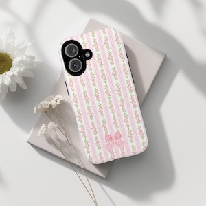 May include: A white phone case with a pink and white floral pattern and a pink bow. The case has a black camera lens.
