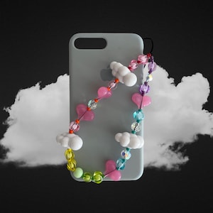 Puede incluir: Una funda de teléfono gris claro con una correa de cuentas de colores. La correa presenta dijes de nubes blancas, dijes de corazones rosas y cuentas azules y verdes.