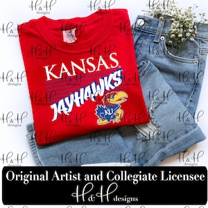 May include: A red short-sleeved t-shirt featuring the white text KANSAS and JAYHAWKS in a bold font, accompanied by a blue and yellow Jayhawk mascot graphic with the letters KU. The shirt is paired with light blue distressed denim jeans.