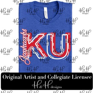 Puede incluir: Una camiseta de manga corta en color azul real con el texto Jayhawks y las letras KU en rojo y blanco con una tipografía estilizada. La prenda tiene cuello redondo y una etiqueta blanca de la marca en el cuello. El diseño está centrado.