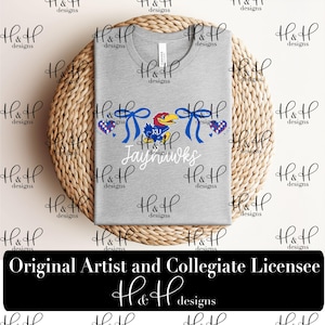 May include: A heather gray crew neck t-shirt featuring a graphic of a blue and red bird mascot, two blue ribbons, two checkered hearts, and the word Jayhawks in white script. The shirt is displayed on a round, woven, light brown textured mat.