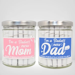 May include: Two glass jars with silver lids filled with rolled up pieces of paper. The jars have pink and blue labels that say "I'm a Badass new Mom" and "I'm a Badass new Dad" with the text "Because I'm absolutely fucking killing it!" underneath.