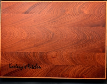 May include: A rectangular wooden cutting board with a natural wood grain finish. The board has a light brown border and is engraved with the text "Kortney's Kitchen".