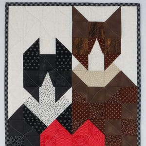 May include: A quilted wall hanging depicting two cat figures, one black and one brown, joined by a red heart. The cats are set against a white background, with a red and white checkered border at the bottom.