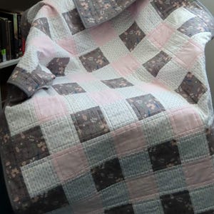 May include: A grey and pink patchwork quilt with a floral pattern. The quilt is folded and draped over a chair.
