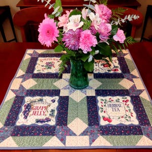 May include: A patterned quilted table runner with floral motifs and labels such as "Grape Jelly" and "Herbal Tea." A green vase holds a bouquet of pink and white flowers, adding a vibrant touch to the scene. The runner is likely 100 cm in length.