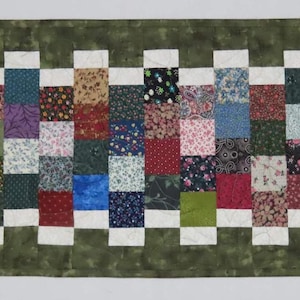 Peut inclure: Un chemin de table matelassé vert avec une bordure à carreaux blancs. Le chemin de table est décoré d'un motif patchwork de carrés colorés.