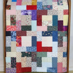 May include: A colourful patchwork quilt featuring a mix of floral, patterned, and solid fabrics. The quilt is displayed on a wooden frame, highlighting its handcrafted quality and vintage style. The quilt's dimensions are approximately 150 cm x 100 cm.