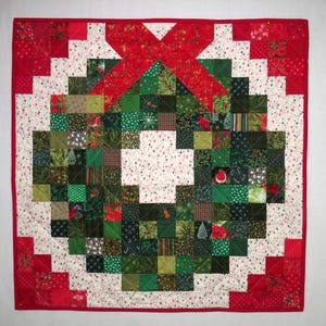 Peut inclure: Un quilt patchwork carré présentant un motif de couronne centrale composé de petits carrés de tissu verts et rouges avec des motifs de fêtes. La couronne est encadrée par une bordure blanche avec des poinsettias rouges et un biais rouge.