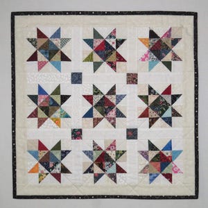 May include: A handmade quilt with nine star patterns in assorted colors and fabrics. The stars are set against a white background, with a black and white dotted border. The quilt is a square shape.