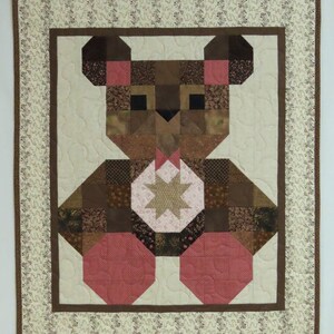 May include: A patchwork quilt with a teddy bear motif. The bear is made from brown and pink fabric pieces, with a star-shaped design on its chest. The quilt has a cream-coloured background and a floral border.
