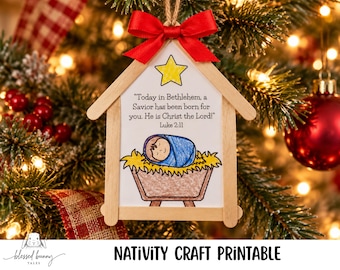 May include: A house-shaped Christmas ornament made of light-colored wooden craft sticks. It features a drawing of a baby in a manger with the text: Today in Bethlehem, a Savior has been born for you. He is Christ the Lord! Luke 2:11. A red bow sits at the top.