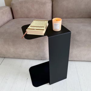 Könnte beinhalten: Ein mattschwarzer, C-förmiger Beistelltisch aus Metall mit einem beigen Buch mit kyrillischem Text und einer brennenden pfirsichfarbenen Kerze in einem Glas mit weißem Etikett, auf dem Superhero Dreams steht.