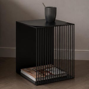 May include: A black metal side table with a solid top and a base featuring vertical rod detailing. The table has a square frame and an open side design, with a dark gray cup placed on the top surface and a book stored on the bottom shelf.