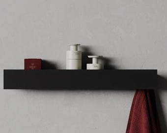 May include: A matte black floating wall shelf holding two white cylindrical containers and a small dark red box. A textured dark red towel hangs from the right side of the shelf. The shelf is mounted horizontally against a plain gray wall.