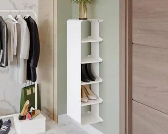 May include: A white, vertical, wall-mounted storage unit with five open shelves. The unit holds black ankle boots and beige pumps on its shelves. A glass vase filled with dark red flowers sits on the top surface of the white storage unit.