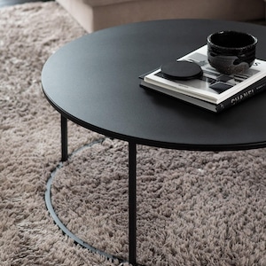 May include: A black round coffee table with a metal base and a black book with a white cover and a black coaster on top. The table is on a light brown shag rug.