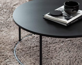 May include: A round black coffee table with a thin metal frame and a circular base. On the tabletop sits a black ceramic bowl, a small black circular coaster, and a hardcover book with the word CHANEL printed on the spine in white letters.