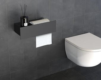 May include: A matte black wall-mounted toilet paper holder with a flat top shelf. The shelf holds a black reed diffuser bottle with black reeds. A roll of white toilet paper is positioned inside the holder, with a sheet hanging down from the front.