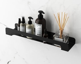 Puede incluir: Un estante de pared de metal negro mate que sostiene tres botellas con dosificador etiquetadas L:A BRUKET, una botella oscura más pequeña y un difusor de varillas de vidrio transparente. El estante tiene una base plana y una barra frontal.