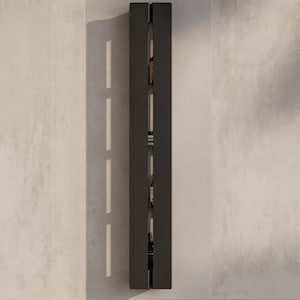 May include: A tall, vertical, matte black wall-mounted storage unit featuring a narrow central opening that reveals multiple interior shelves. Several small bottles are arranged on the shelves within the recessed vertical channel of the unit.
